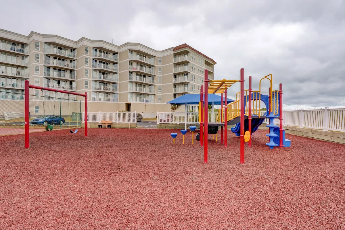 A colorful playground with swings and play structures sits outdoors, inviting children to play while families gather around. The joyful scene radiates community energy, with green grass underfoot and a bright sky overhead.