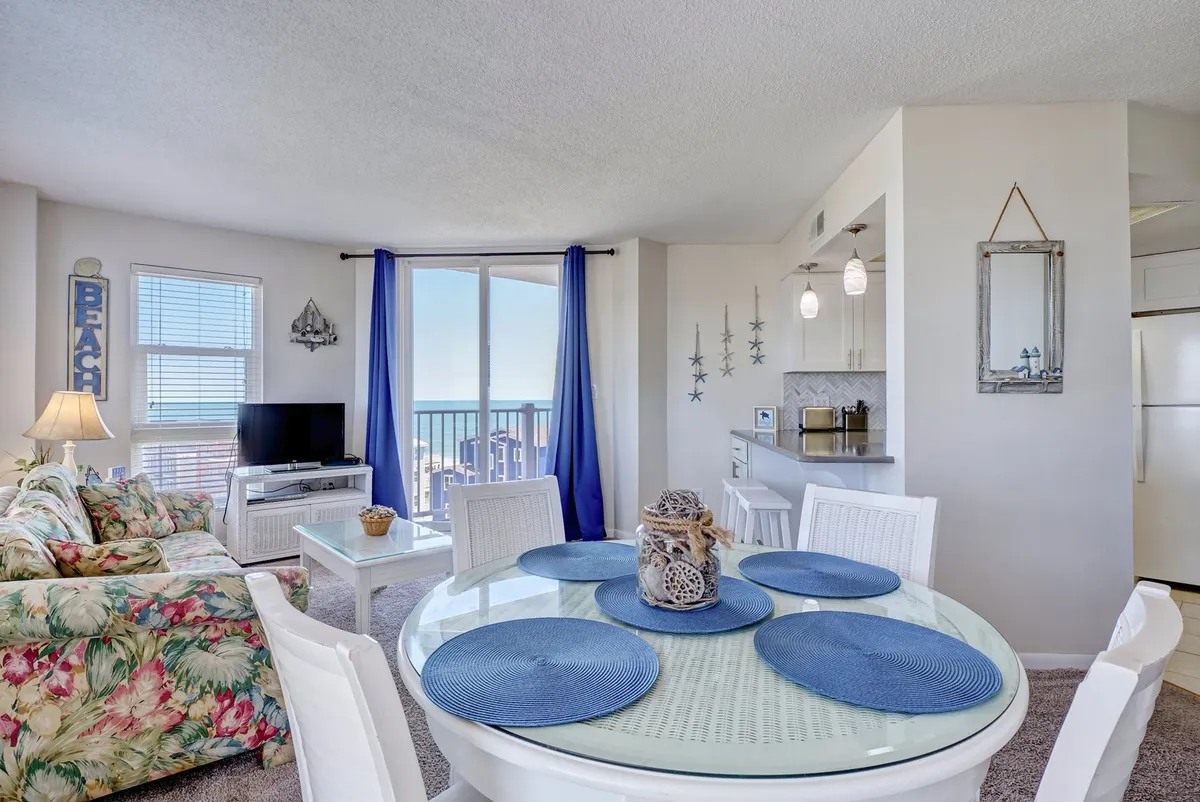A spacious living area with a comfortable floral-patterned couch, blue-accented dining table, and TV, all arranged to maximize enjoyment of the ocean views visible through large windows. Coastal décor and plenty of natural light create a welcoming, cheerful setting ideal for gatherings, meals, and relaxation.