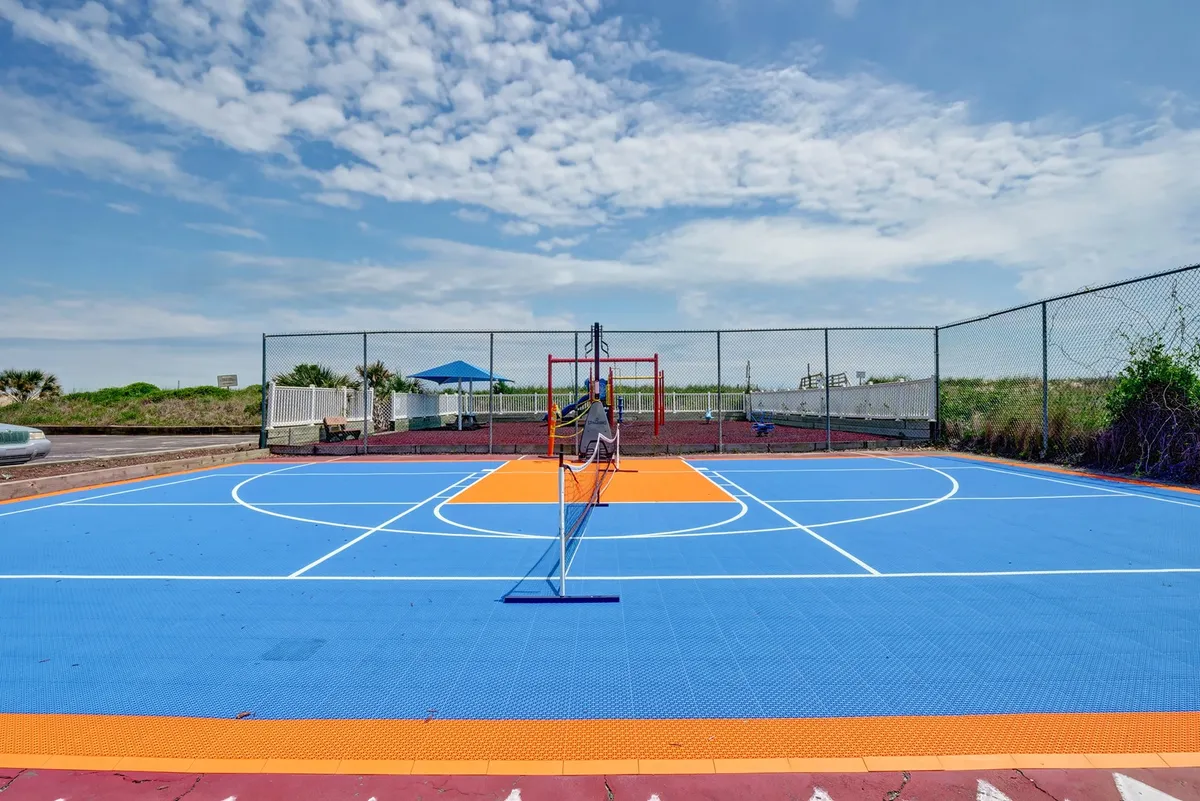 An exterior view of a vibrant blue and orange sports court, featuring a tennis net with playground equipment visible in the background. The scene suggests a multi-use recreational space ideal for diverse activities and community engagement.
