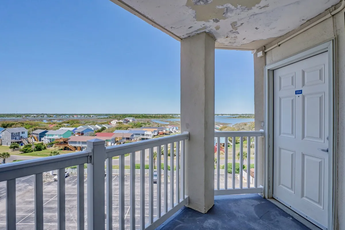 A private balcony with a sturdy railing overlooks tranquil water and surrounding homes. The scene is set for enjoying morning coffee, soaking up sunrise and sunset views, or gathering with friends to feel the salt air and watch the waves. The open doorway visually connects the balcony to the indoor living space.