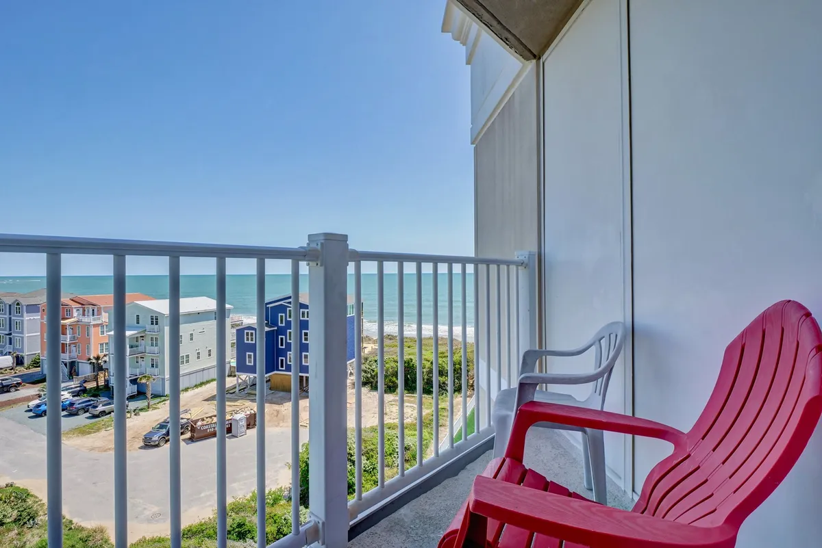 An inviting red Adirondack chair sits on a breezy patio, overlooking the sandy beach and colorful houses along the shoreline. The ocean horizon glows in the soft morning or evening light, suggesting a peaceful spot for sipping coffee or sharing a sunset cocktail.