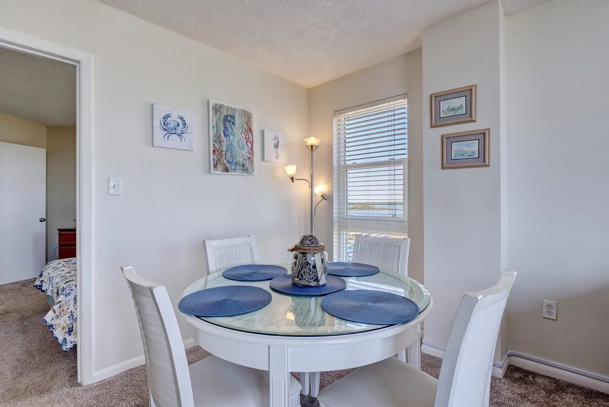 A cozy dining area, softly lit by natural sunlight, features an elegantly set table surrounded by decorative accents. A large window offers glimpses of the outdoors, making the space feel open and inviting—perfect for intimate dinners or quiet moments together.