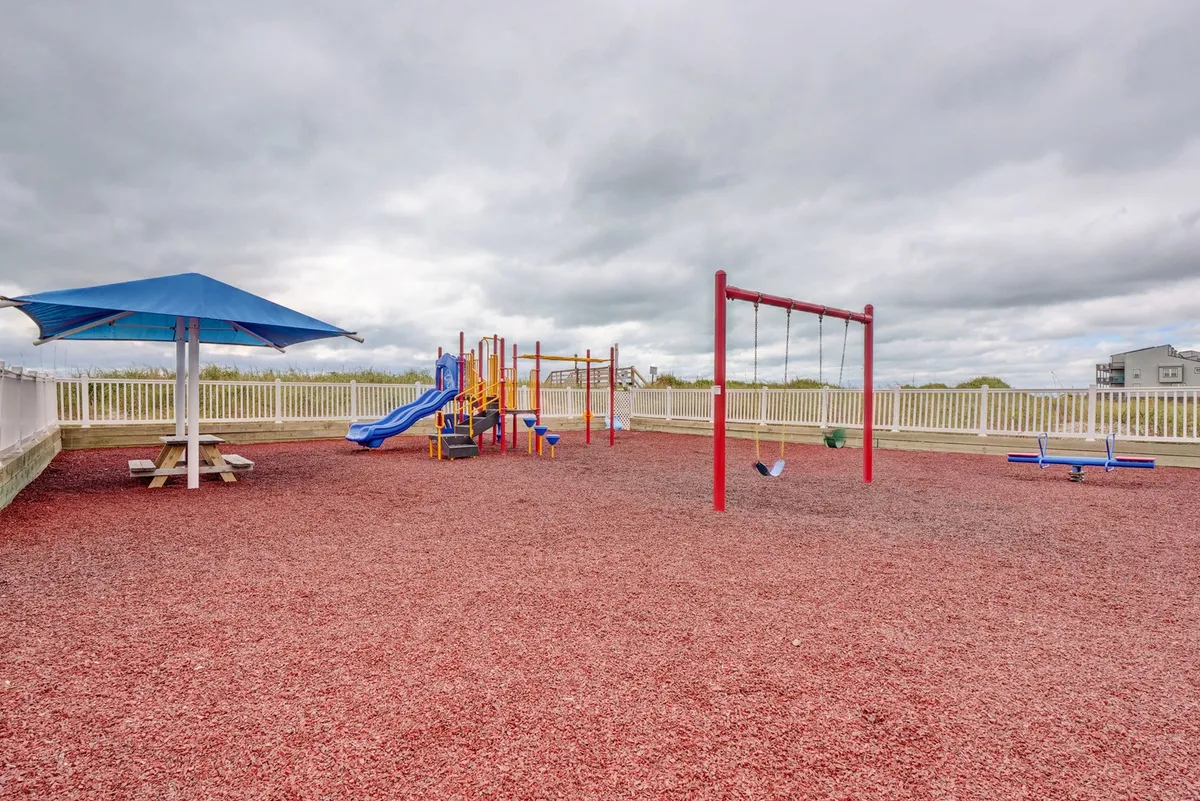 A lively exterior playground area featuring colorful equipment, swings, a picnic space under a shade structure, and a backdrop of families enjoying the outdoors. The environment exudes fun, community, and family-friendly charm.