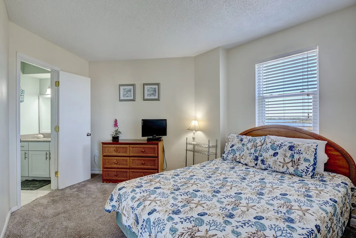 This image features a master bedroom with a bed covered in floral bedding, a wooden dresser, TV, lamp, and framed pictures on the wall, creating a tranquil and inviting atmosphere.