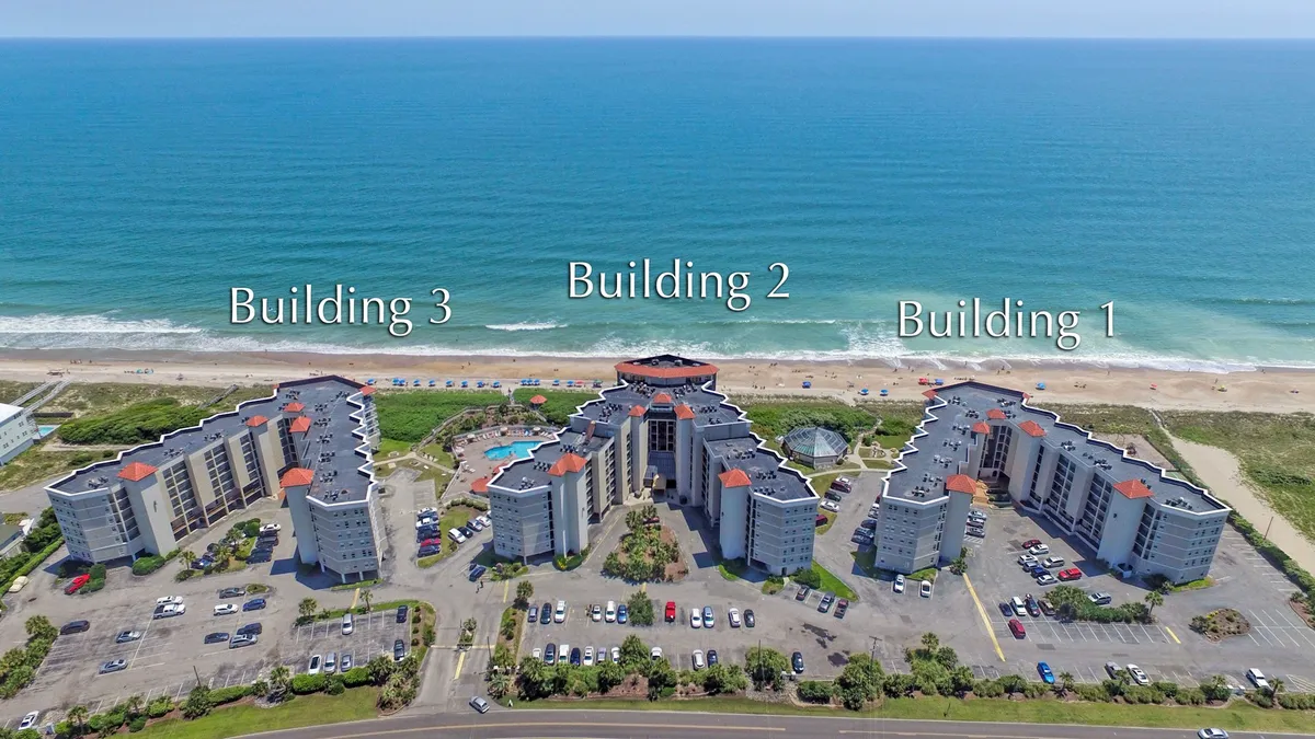 A sweeping exterior view captures three resort buildings framing a sparkling swimming pool, with the Atlantic Ocean stretching beyond. Balconies overlook both the pool and the ocean, reflecting the coastal setting and resort atmosphere. The scene is lively yet serene, hinting at direct beach access and the promise of oceanfront relaxation.