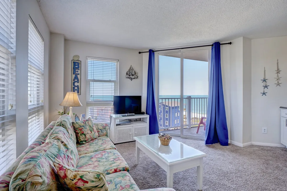 A bright and cheerful living room with a floral-patterned couch positioned to face both a TV and expansive glass sliding doors that open to an unobstructed ocean view. Beach-themed décor accents reinforce the coastal vibe, and sunlight streams through the windows, illuminating the inviting seating area.