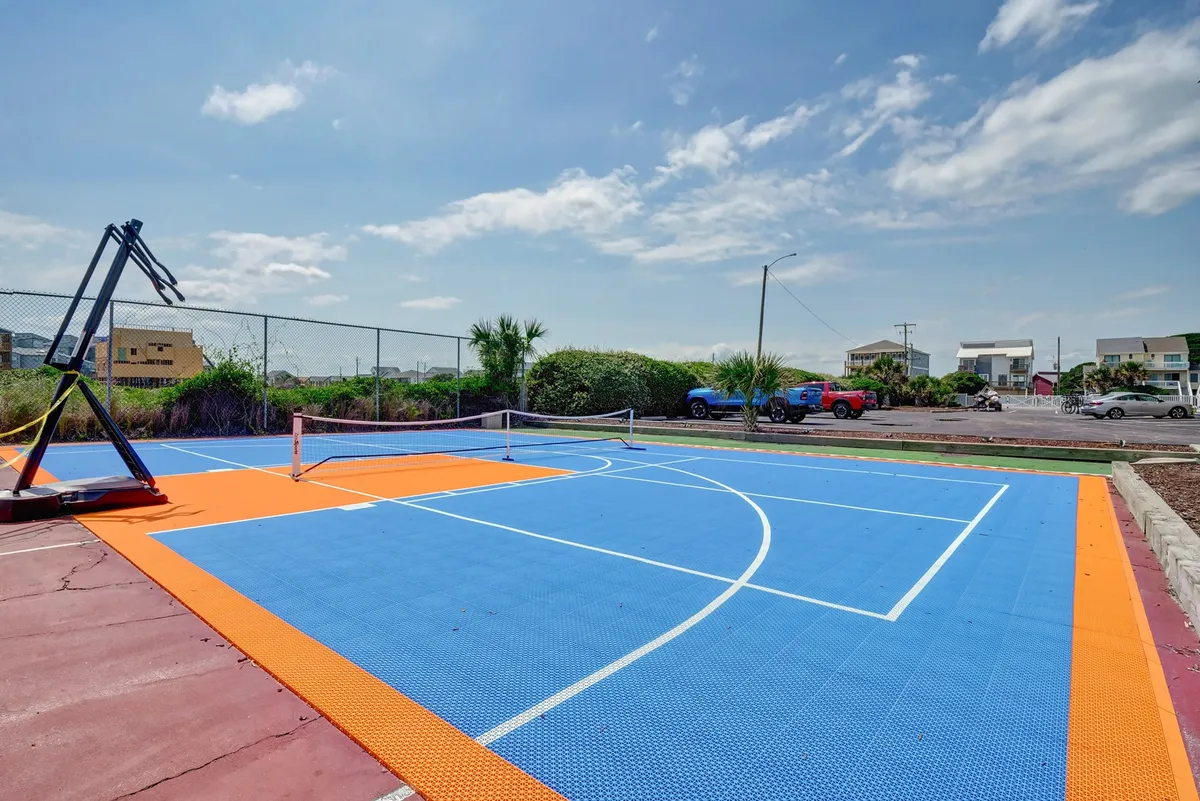 An outdoor basketball court with a tennis net, surrounded by greenery and an open sky. The court surface is clean and well-marked, and the area is bathed in natural light, suggesting an inviting space for both casual and organized sports.